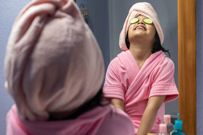 A cute indian girl child in pink bathrobe applying cucumber slices to her eyes in front of mirror