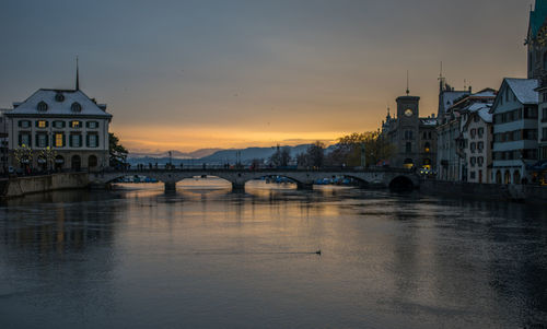 Bridge over river by buildings against sky during sunset