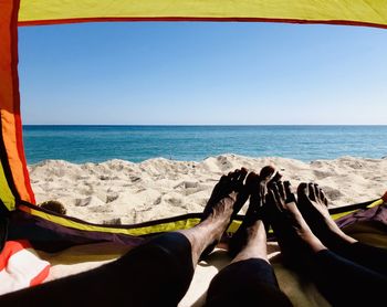 Low section of couple relaxing in tent on beach at sea against sky during sunny day