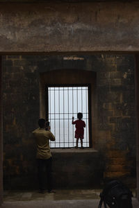 Father photographing son while standing at window