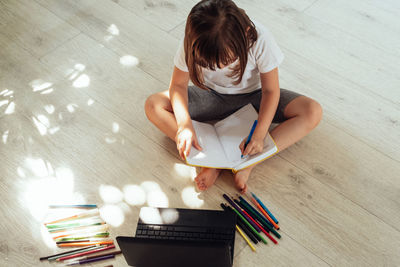 Child sits on the floor with a laptop, pen, notepad and colored pencils. 