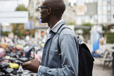 Side view of mid adult businessman looking away while standing with smart phone in city