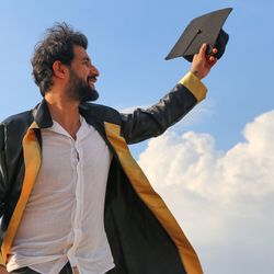 Man in graduation gown holding mortarboard against sky