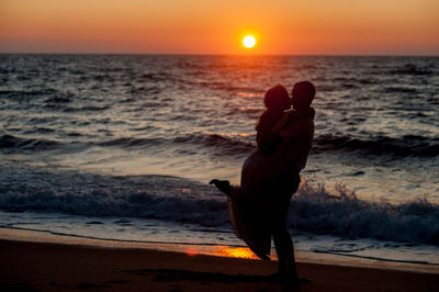 Side view of silhouette man kissing woman at beach against sky during sunset