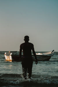 Rear view of man walking in sea towards his boat against clear sky