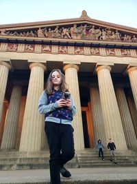 Full length portrait of girl holding camera while standing against historic building