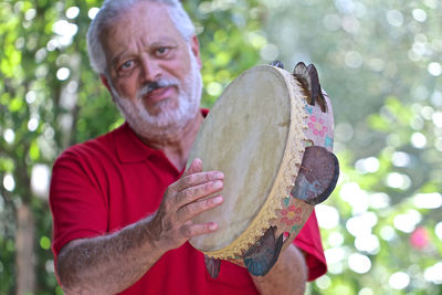 Portrait of man playing tambourine while standing against trees