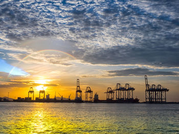 Silhouette cranes at harbor against sky during sunset