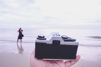 Cropped hand with camera at beach against sky