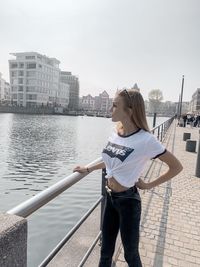 Man standing on railing by river in city against sky