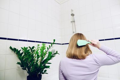 Rear view of woman holding hairbrush against wall in bathroom