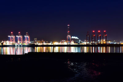 Illuminated factory by sea against clear sky at night