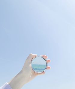 Cropped hand of person holding mirror against clear blue sky