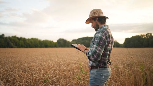 Bearded man in checkered shirt and hat browsing tablet while checking cereal crops