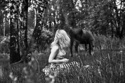 Young blonde woman sitting in the forest grass looking back at a grazing horse. black and white.