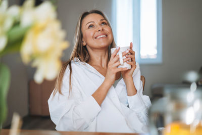 Young happy woman with blonde hair in white shirt drinking tea with bouquet on dinner table at home