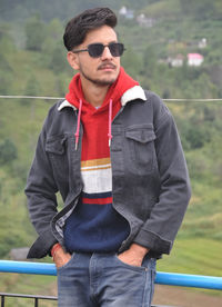 A indian young guy wearing sunglasses posing with his hands on pocket, standing outdoor 