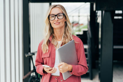 Happy blond woman in pink jacket holding laptop computer