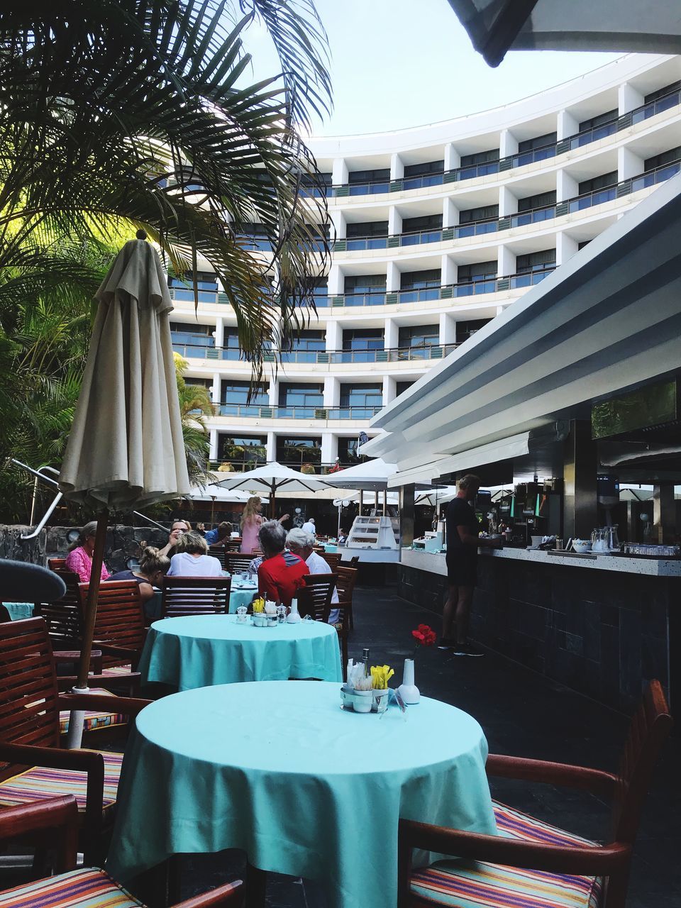 table, architecture, restaurant, built structure, seat, chair, cafe, day, building exterior, business, tree, real people, plant, incidental people, food and drink, palm tree, outdoors, tropical climate, group of people, people, setting