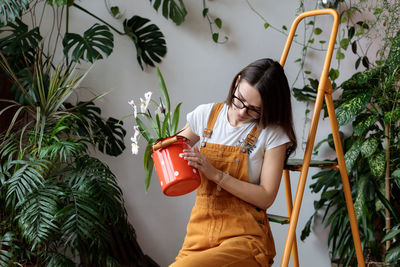 Young woman at home garden, female looking at orchid flower in can, sitting on ladder in plant shop