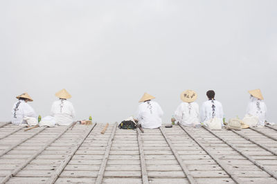 Rear view of people wearing asian style conical hats sitting in row against sky