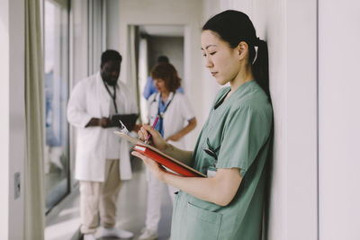 Side view of female doctor writing on clipboard in hospital corridor