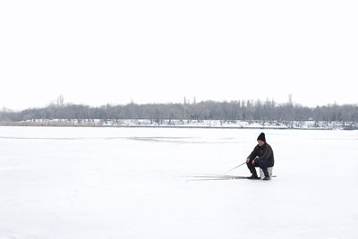 Full length of man fishing on frozen lake against clear sky