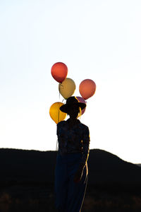 Silhouette of unrecognizable female in hat standing with heap of colorful balloons against cloudless sky at sunset time against mountain ridge