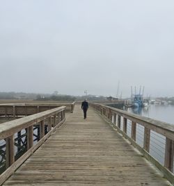 Rear view of man walking on pier over sea against sky during foggy weather