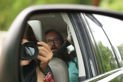 Reflection of woman photographing by man sitting in side-view mirror of car