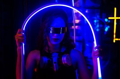 Portrait of young woman wearing sunglasses while standing against illuminated nightclub