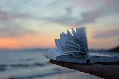 Cropped image of hand holding book against sea during sunset