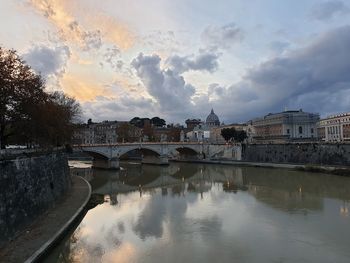 Bridge over river by buildings against sky during sunset