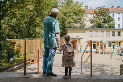 Senior man with granddaughter standing near railing at kindergarten