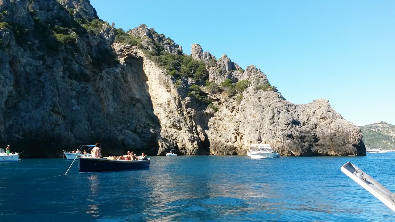 water, sea, mountain, waterfront, nautical vessel, clear sky, scenics, tranquil scene, beauty in nature, blue, tranquility, rock formation, transportation, nature, cliff, rock - object, mode of transport, boat, idyllic, rocky mountains