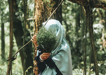 Woman in hijab covering face with plants in forest