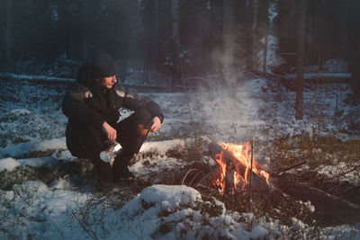 Full length of man sitting by campfire in forest during winter