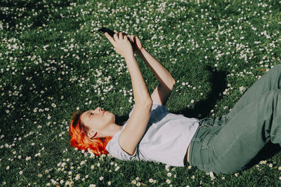 Side view of delighted redhead female lying on grass on lawn and messaging on mobile phone in summer
