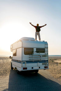 Back view of carefree male explorer standing on roof of camper parked on seashore and enjoying freedom with outstretched arms against sundown sky