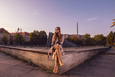 Young woman sitting on retaining wall against sky