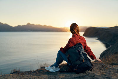 Rear view of woman sitting on shore against sky during sunset