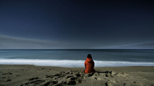 Rear view of woman sitting at beach against sky at night