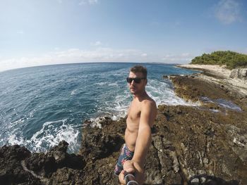 Portrait of shirtless man standing on rocky shore against sky