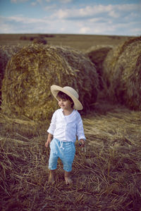 Boy a child in a straw hat and blue pants stands in a mowed field with stacks in the summer