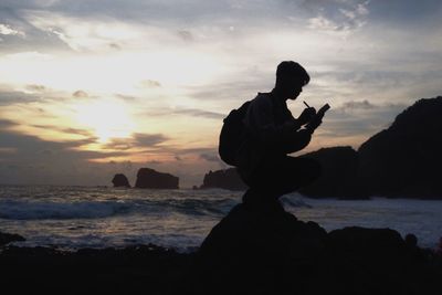 Silhouette man crouching on rock at beach during sunset