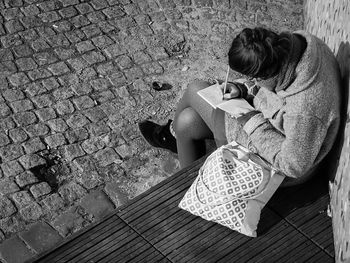 High angle view of woman sitting on footpath writing in a paper notebook 