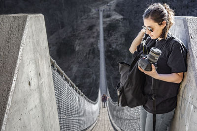 Backpacker holding digital camera while standing on footbridge