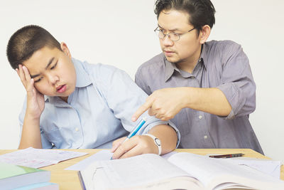 Frustrated father teaching son while sitting at table against white background