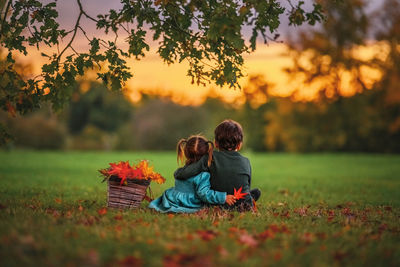 Siblings are watching the autumn sunset sitting in the grass under a tree