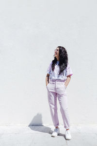 Smiling young woman with hands in pockets looking away while standing in front of white wall